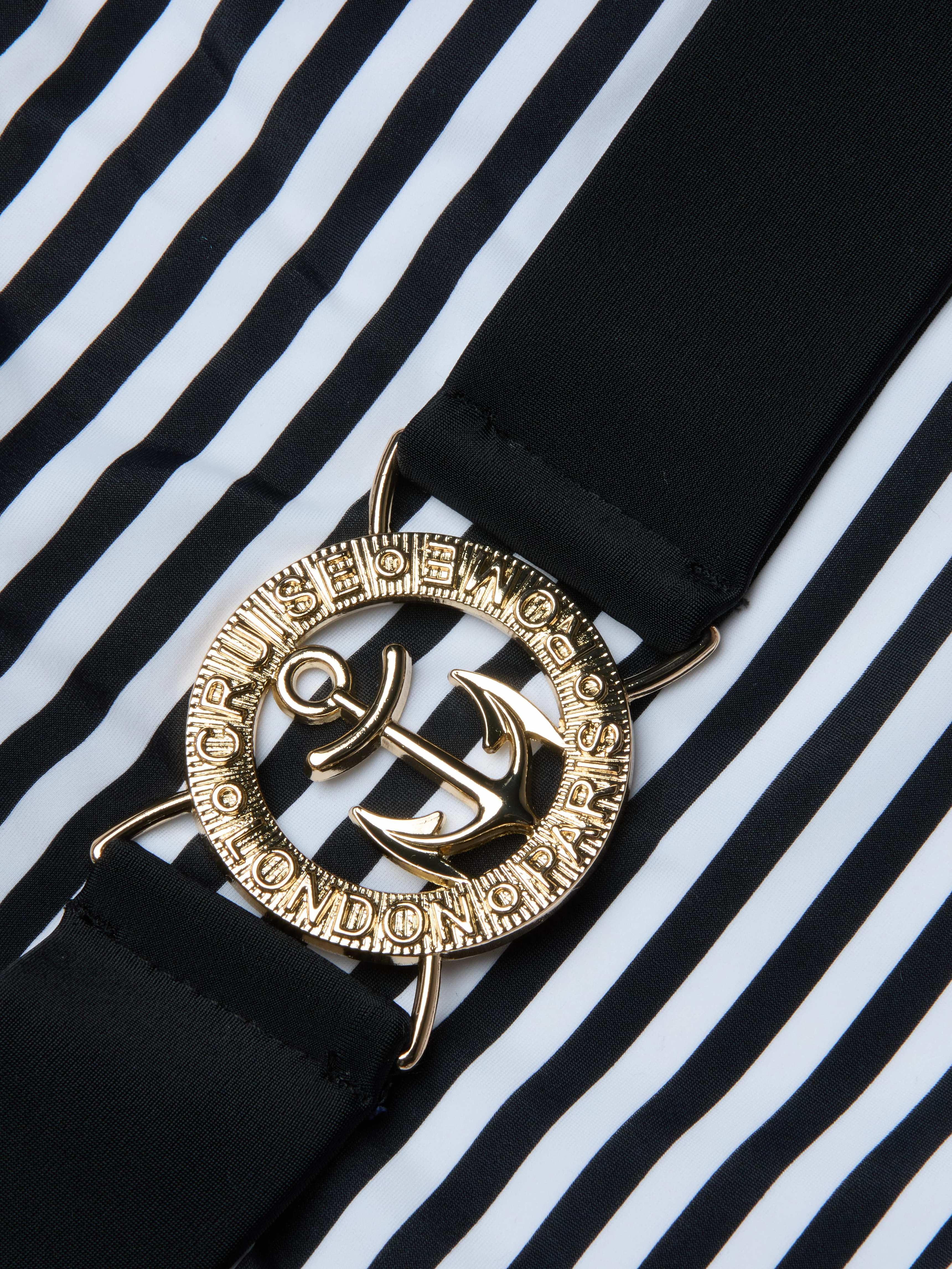 A close-up of a gold anchor belt buckle with engraved text, attached to a black strap, set against the bold black and white vertical stripes of the Mila One-Piece Swimsuit.