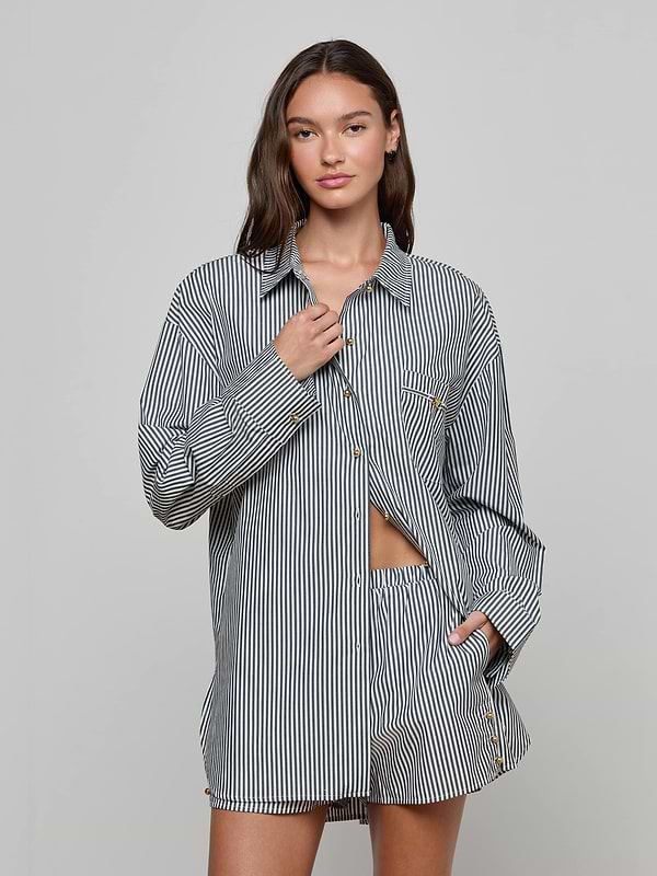 A woman stands against a plain background wearing the Lorena Cover-Up Shirt, a long-sleeved black-and-white striped resort top with gold buttons, paired with matching shorts. She has one hand at her collar and the other in her pocket, looking confidently at the camera.