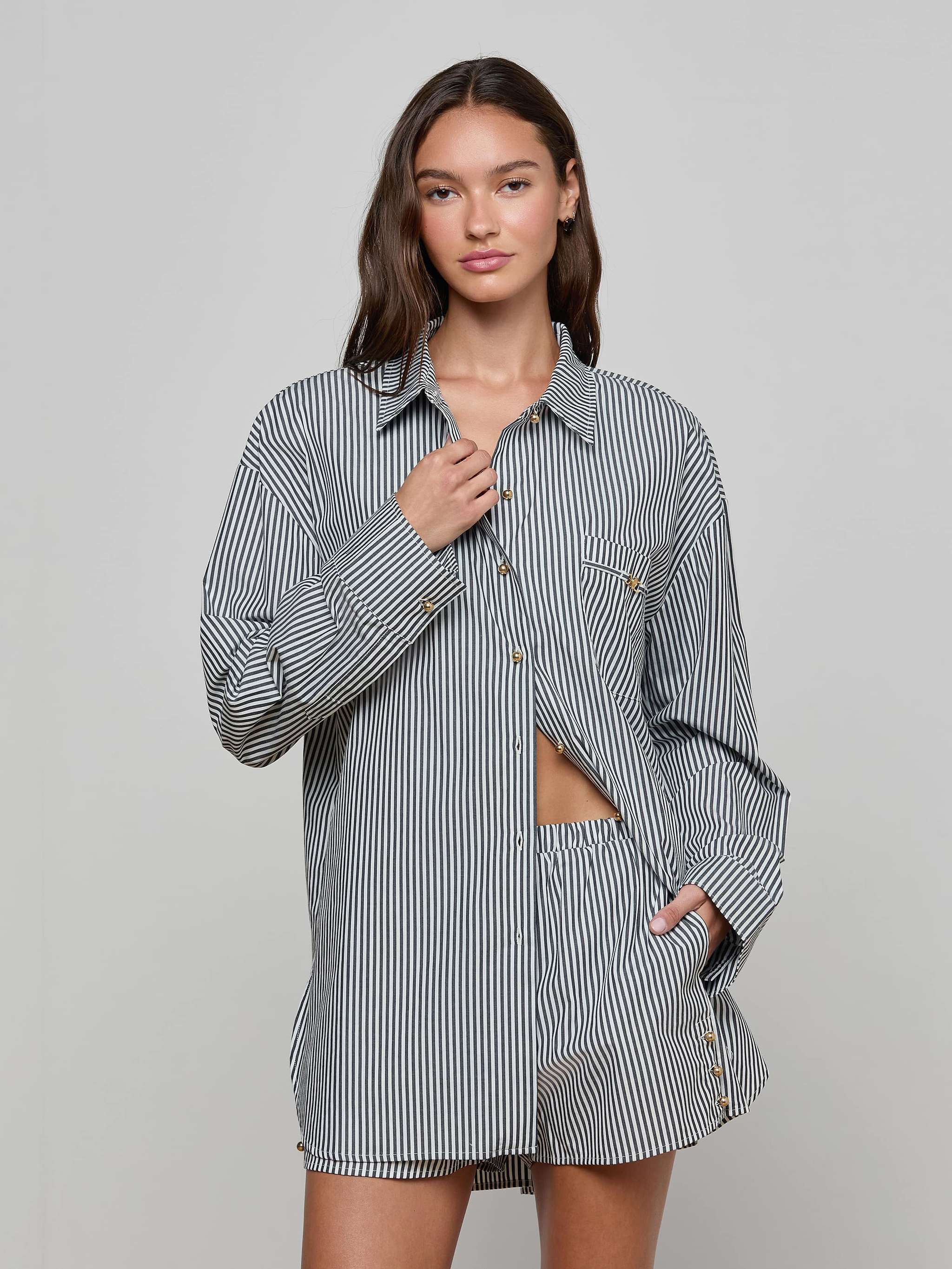 A woman stands against a plain background wearing the Lorena Cover-Up Shirt, a long-sleeved black-and-white striped resort top with gold buttons, paired with matching shorts. She has one hand at her collar and the other in her pocket, looking confidently at the camera.
