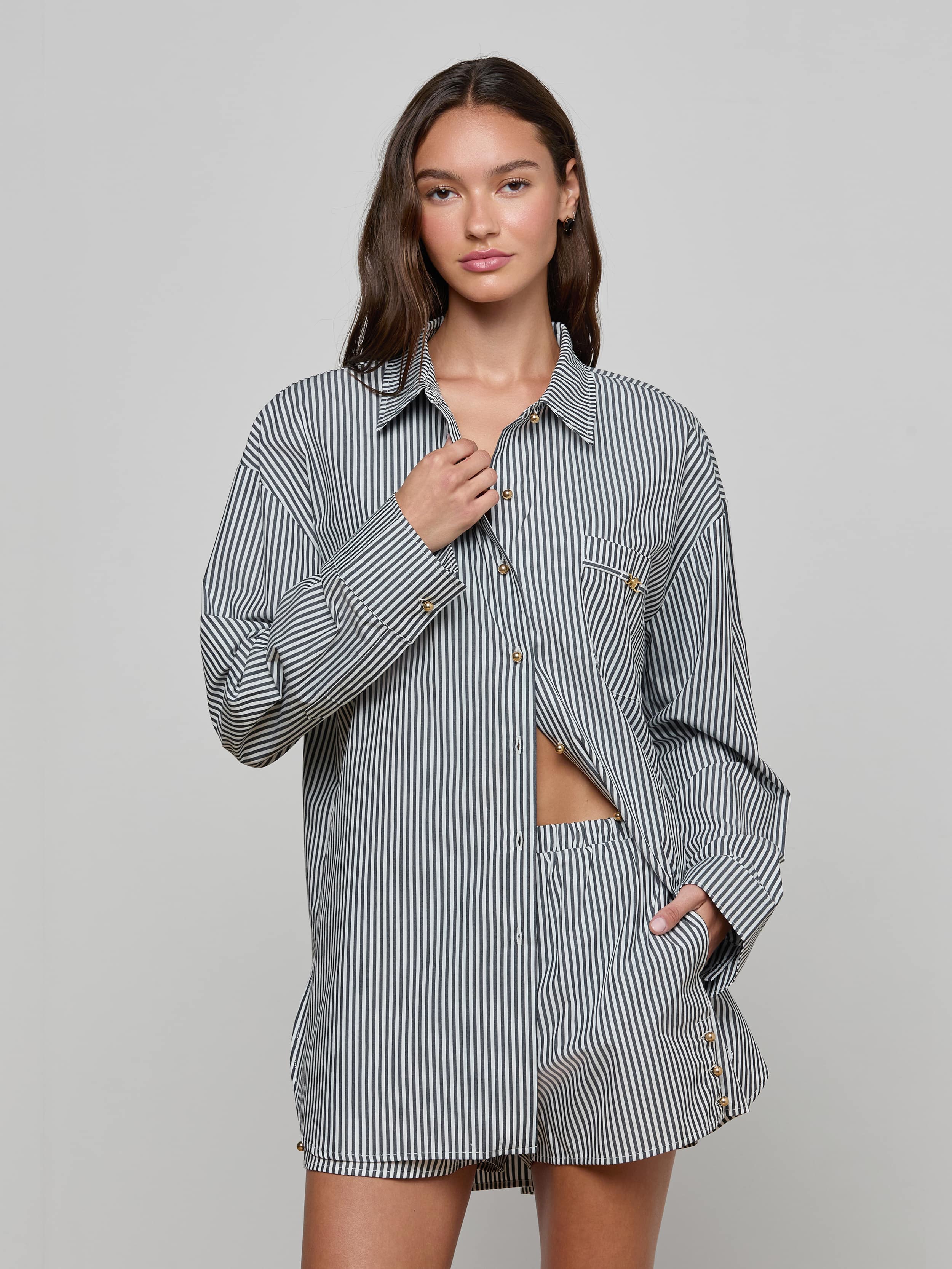 A woman stands against a plain background wearing the Lorena Cover-Up Shirt, a long-sleeved black-and-white striped resort top with gold buttons, paired with matching shorts. She has one hand at her collar and the other in her pocket, looking confidently at the camera.