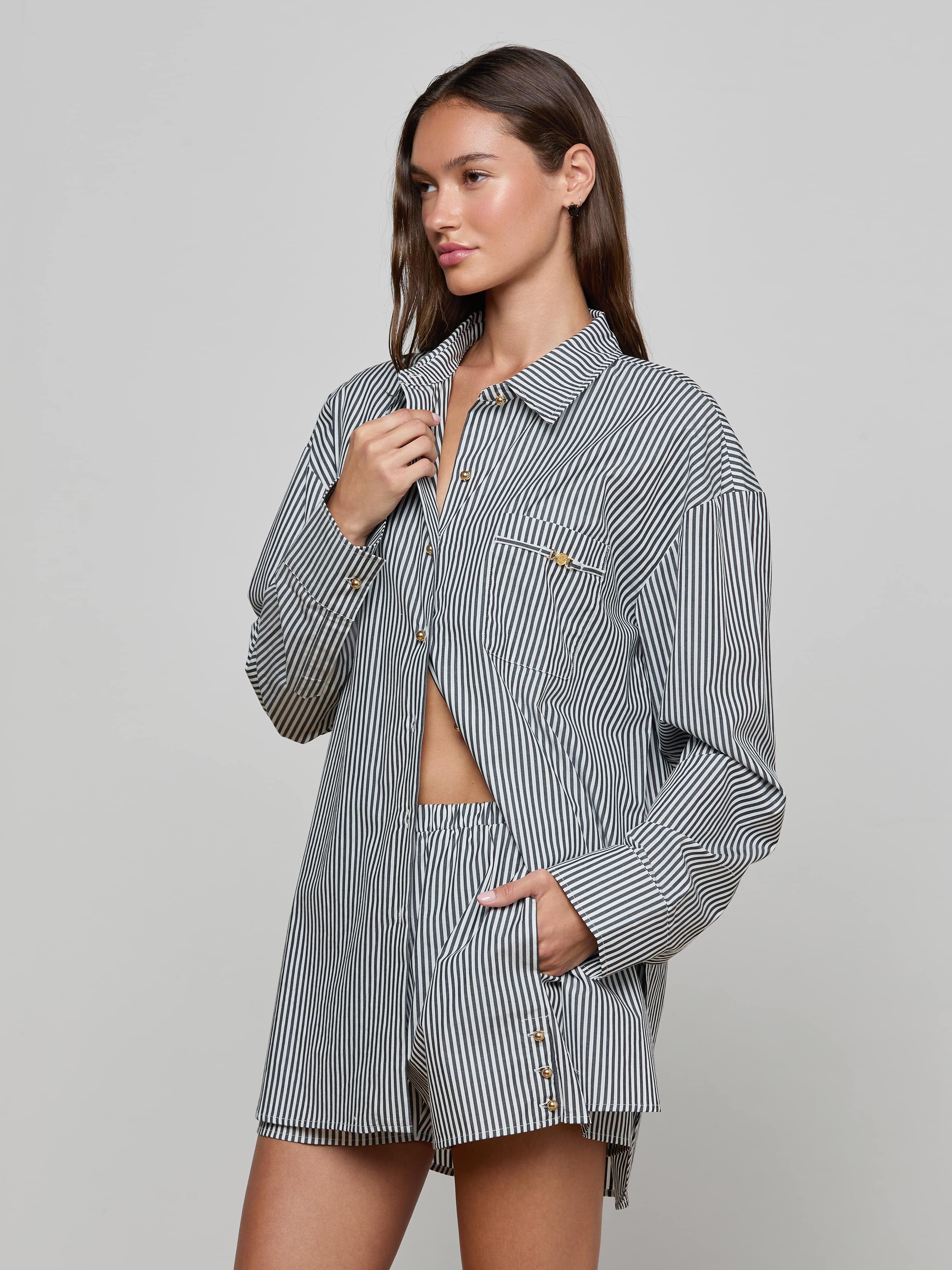 A woman wearing the Lorena Cover-Up Shirt, a loose, long-sleeve resort top in black-and-white striped cotton poplin with matching shorts, stands against a plain light background—one hand holding the shirt collar and the other tucked into a pocket.