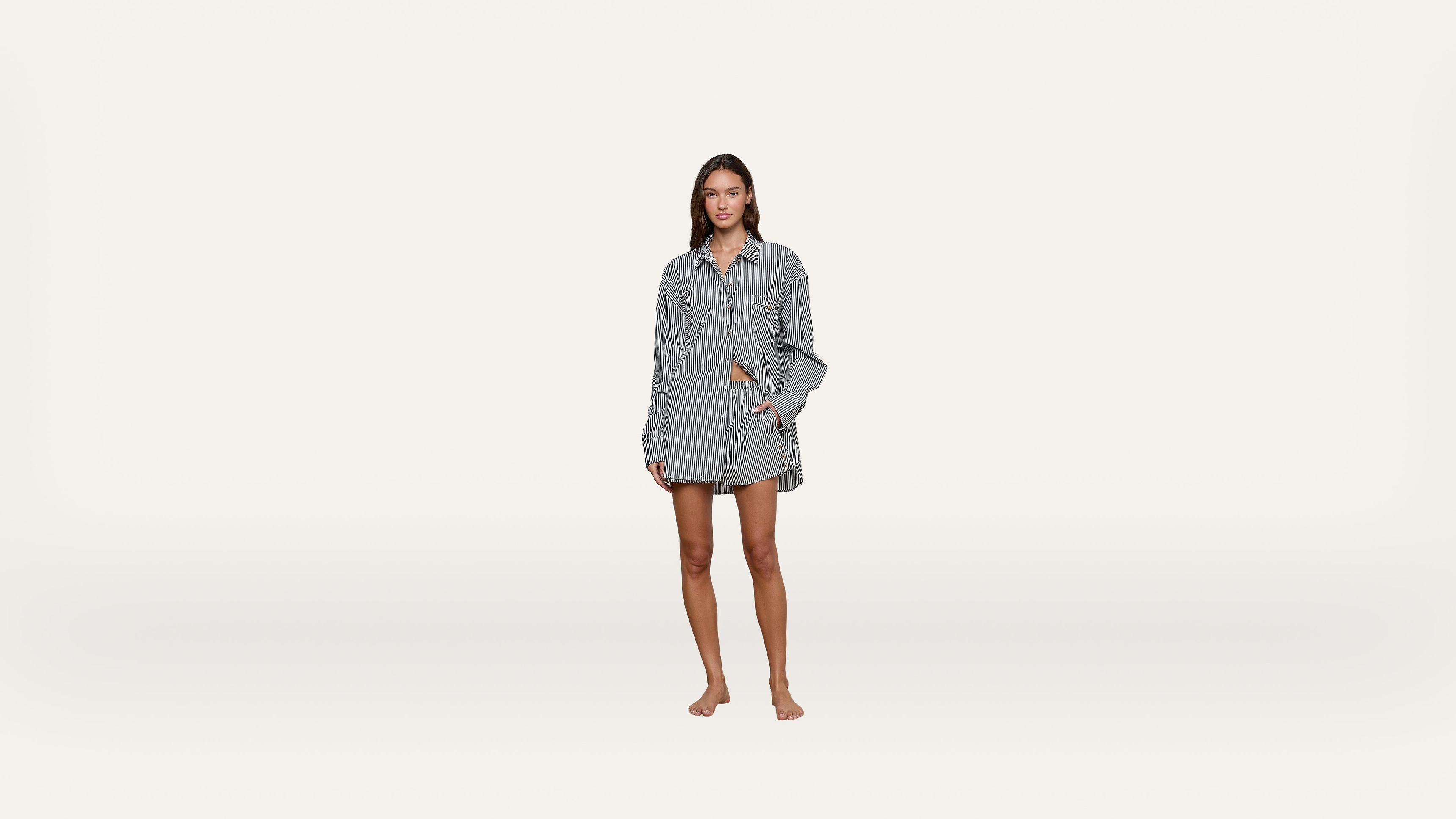 A woman stands barefoot wearing a loose, long-sleeve, button-up shirt and matching shorts. The background is plain white, and she is looking at the camera with a relaxed pose.