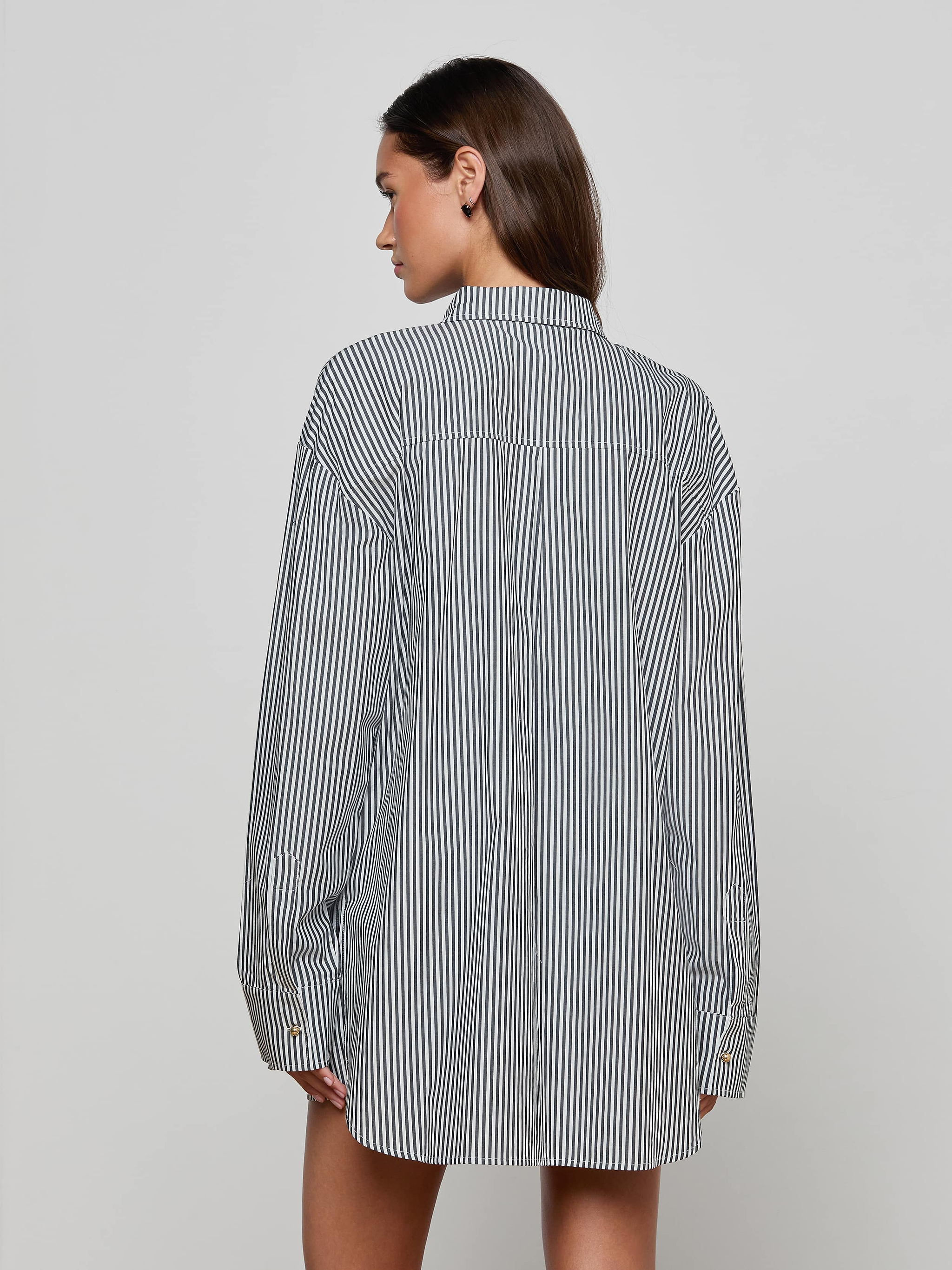 A woman with straight brown hair is standing with her back to the camera against a plain, light background, wearing the Lorena Cover-Up Shirt—a long-sleeve, black-and-white striped resort style.