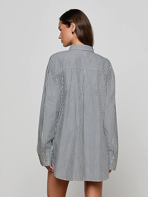 A person with long brown hair is shown from the back, wearing the Lorena Cover-Up Shirt—an oversized black and white vertically striped cotton poplin long-sleeve shirt—against a plain light gray background.