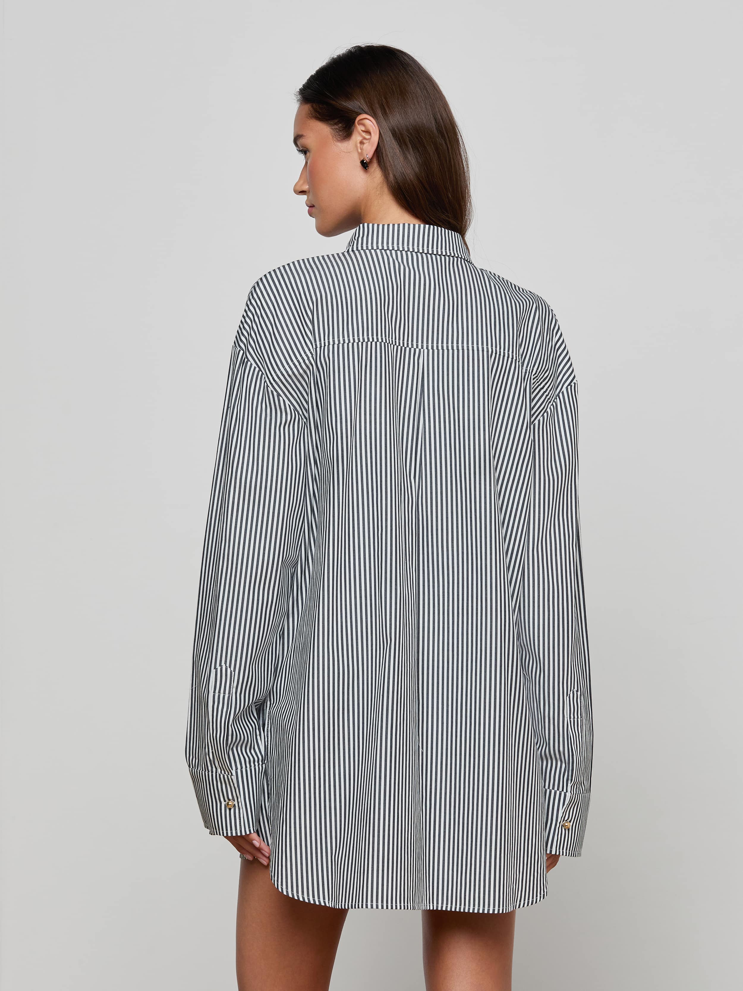 A woman with straight brown hair is standing with her back to the camera against a plain, light background, wearing the Lorena Cover-Up Shirt—a long-sleeve, black-and-white striped resort style.