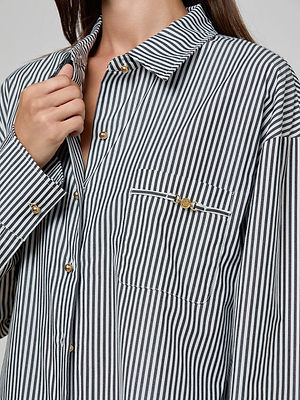 A person wearing the Lorena Cover-Up Shirt—a black and white vertically striped cotton poplin piece with gold buttons, a button-flap front pocket, and resort collection detailing—holds the collar with one hand.