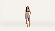 A woman wearing a black bikini top and gray drawstring shorts stands barefoot against a plain white background, looking at the camera with one hand in her pocket.
