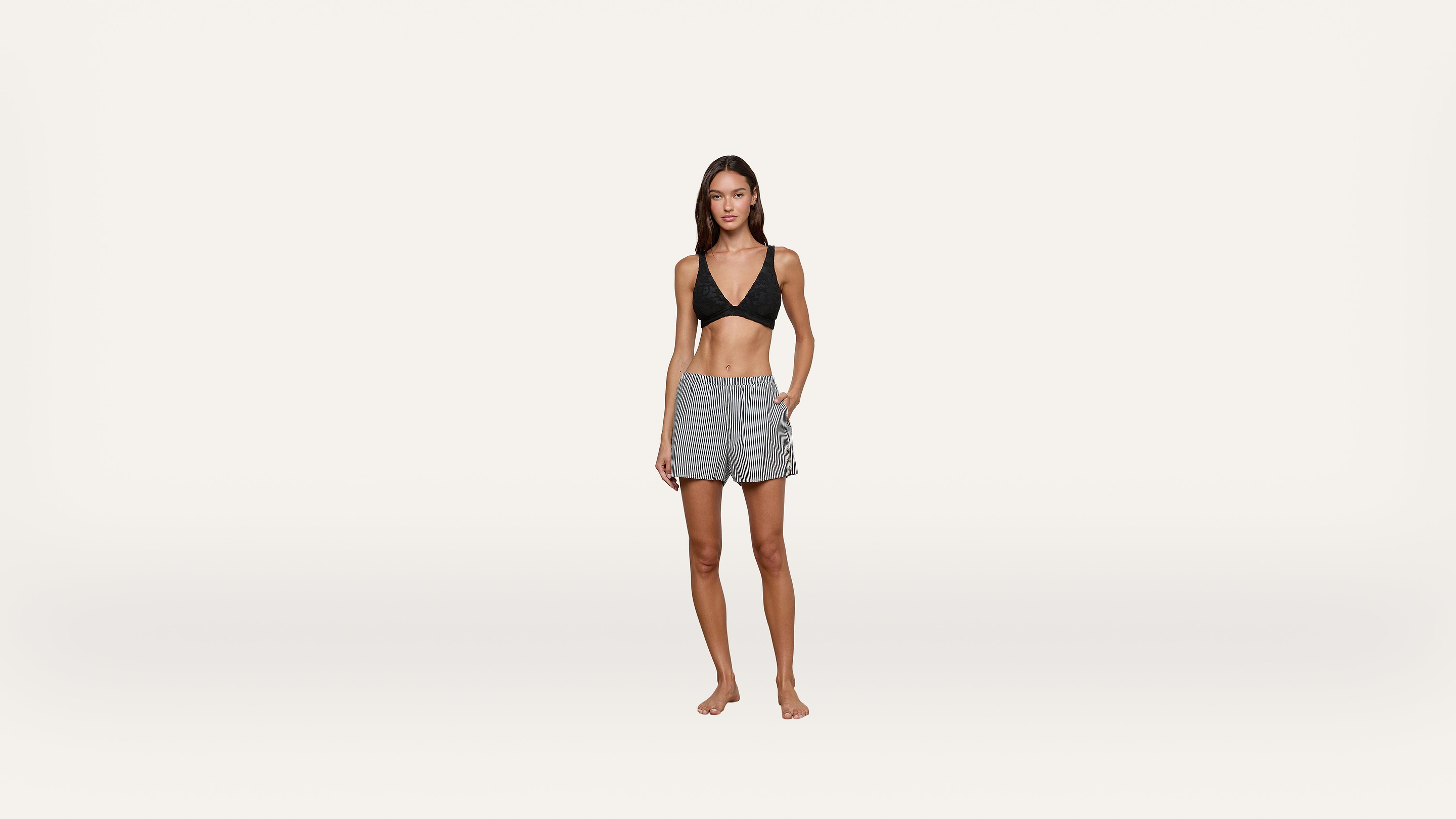 A woman wearing a black bikini top and gray drawstring shorts stands barefoot against a plain white background, looking at the camera with one hand in her pocket.