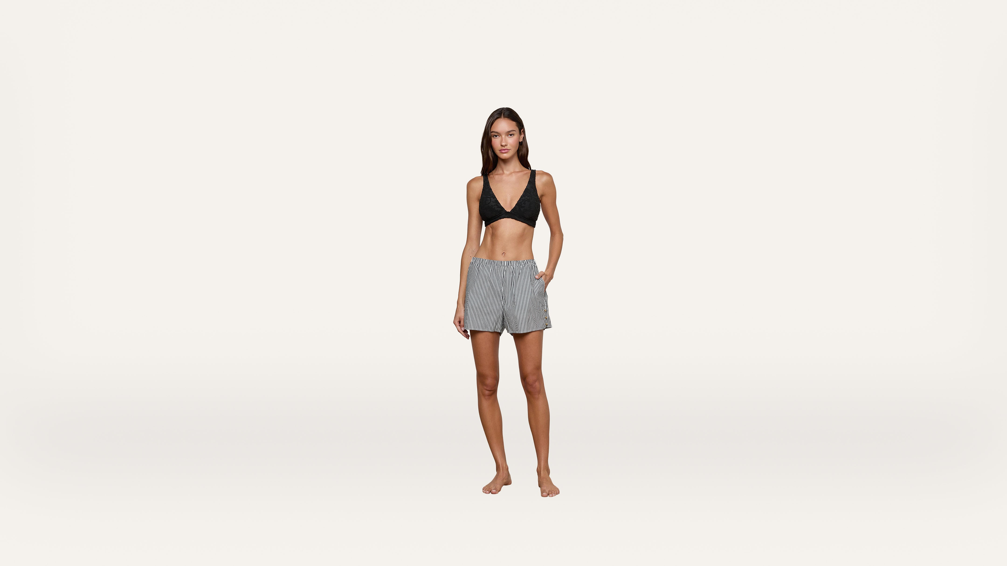 A woman wearing a black bikini top and gray drawstring shorts stands barefoot against a plain white background, looking at the camera with one hand in her pocket.