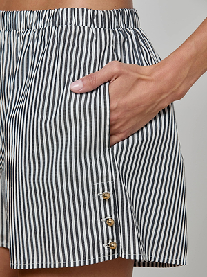Close-up of a person wearing the Amiya Cover-Up Short—a tailored black and white vertically striped cotton poplin style with gold buttons near the hem; their hand rests in the pocket.