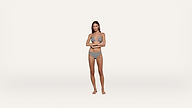A woman stands barefoot on a white background, wearing a black and white striped bikini, with her arms folded across her stomach and a neutral expression.