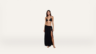 A woman stands barefoot on a plain background, wearing a black bikini top and a long black skirt with a thigh-high slit.