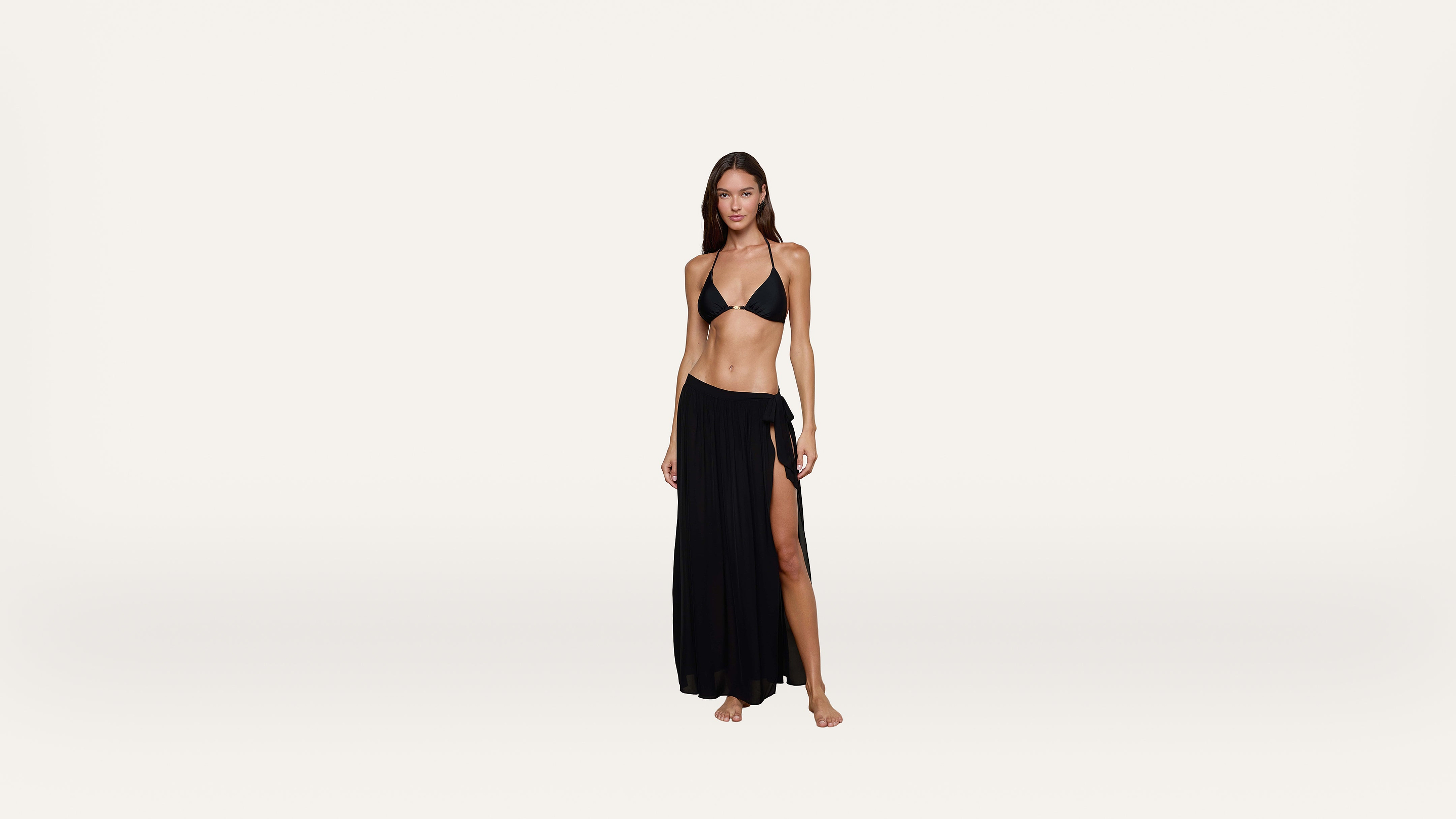 A woman stands barefoot on a plain background, wearing a black bikini top and a long black skirt with a thigh-high slit.