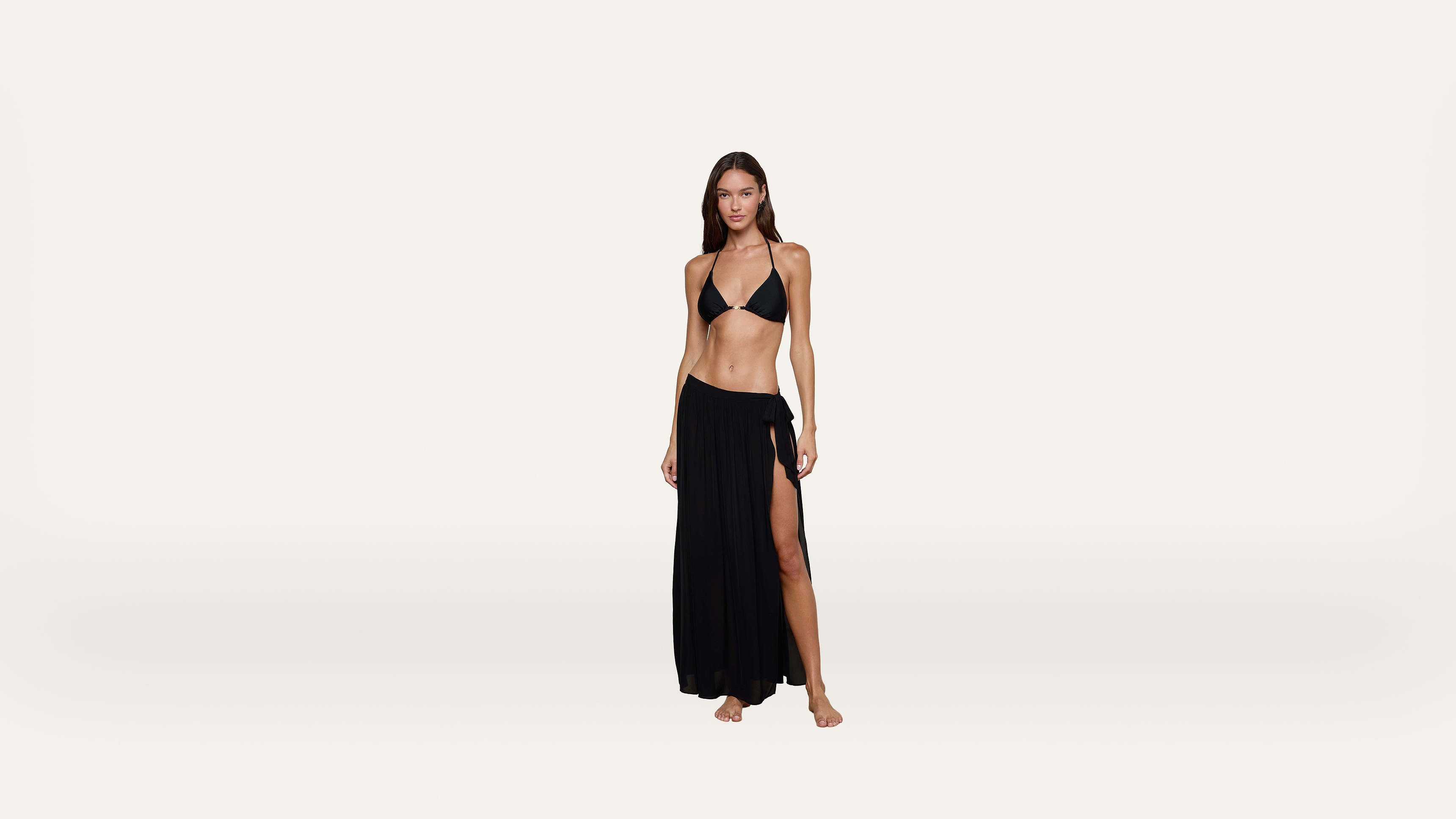 A woman stands barefoot on a plain background, wearing a black bikini top and a long black skirt with a thigh-high slit.