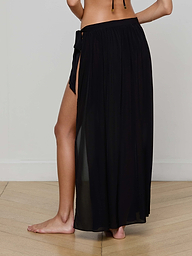 A person wearing the Klaire Cover-Up Skirt—a black sheer viscose chiffon wrap maxi skirt with a high side slit and tie closure—stands barefoot against a plain light background.