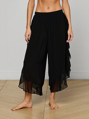 A person stands barefoot on a wooden floor, wearing the Adonis Cover-Up Pant in black, made from sheer viscose chiffon. The image is cropped at the shoulders and ankles with a plain light-colored wall as the background.