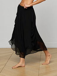 A person wears the Adonis Cover-Up Pant, a flowing black sheer chiffon midi skirt with an elastic waistband, while walking barefoot against a light background. Only the lower torso and legs are visible.