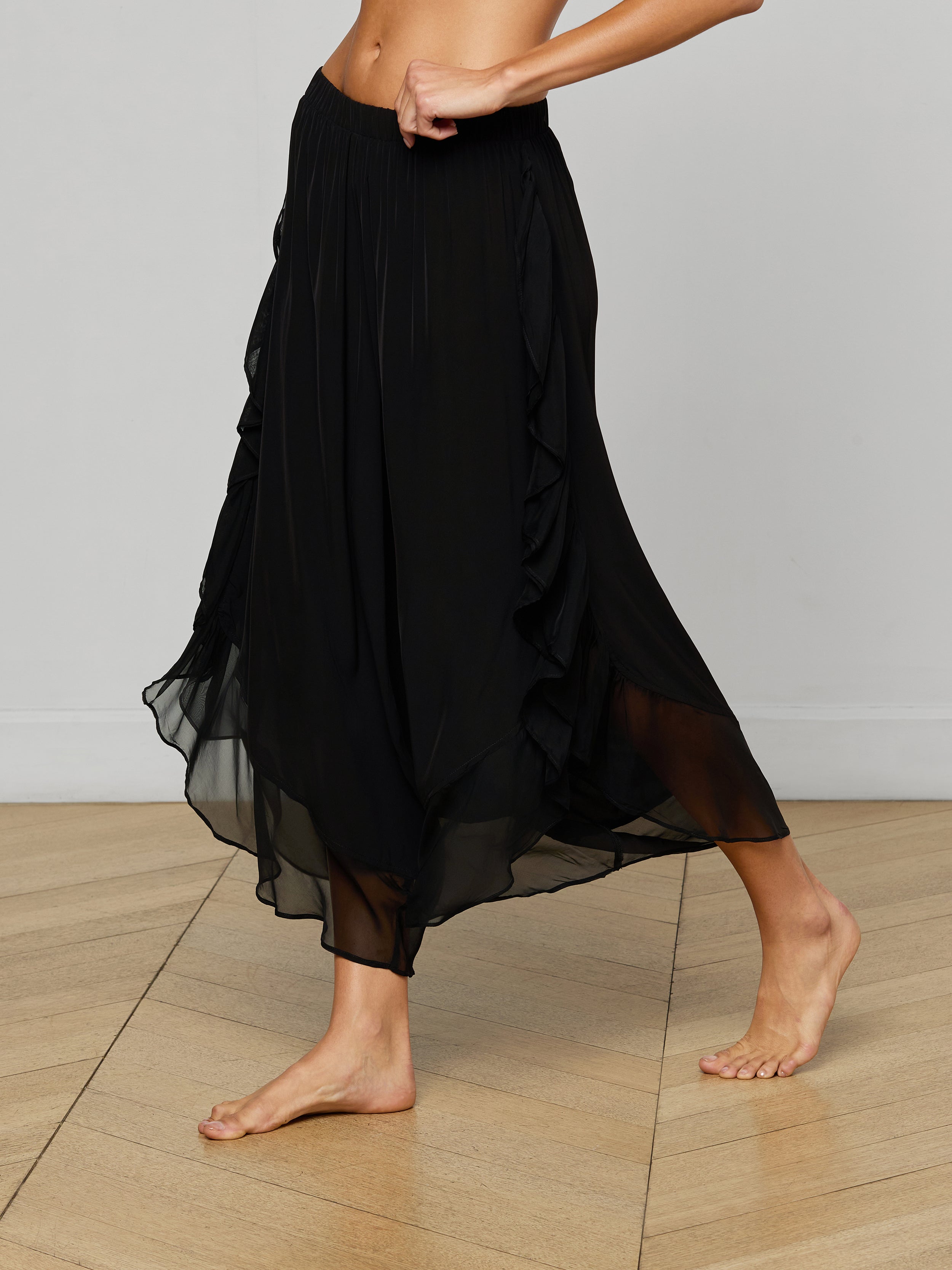 A person wears the Adonis Cover-Up Pant, a flowing black sheer chiffon midi skirt with an elastic waistband, while walking barefoot against a light background. Only the lower torso and legs are visible.