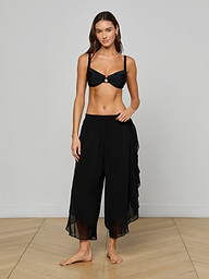 A woman stands barefoot against a plain light background, wearing a black bikini top and the Adonis Cover-Up Pant—loose, flowy pants made from sheer chiffon.