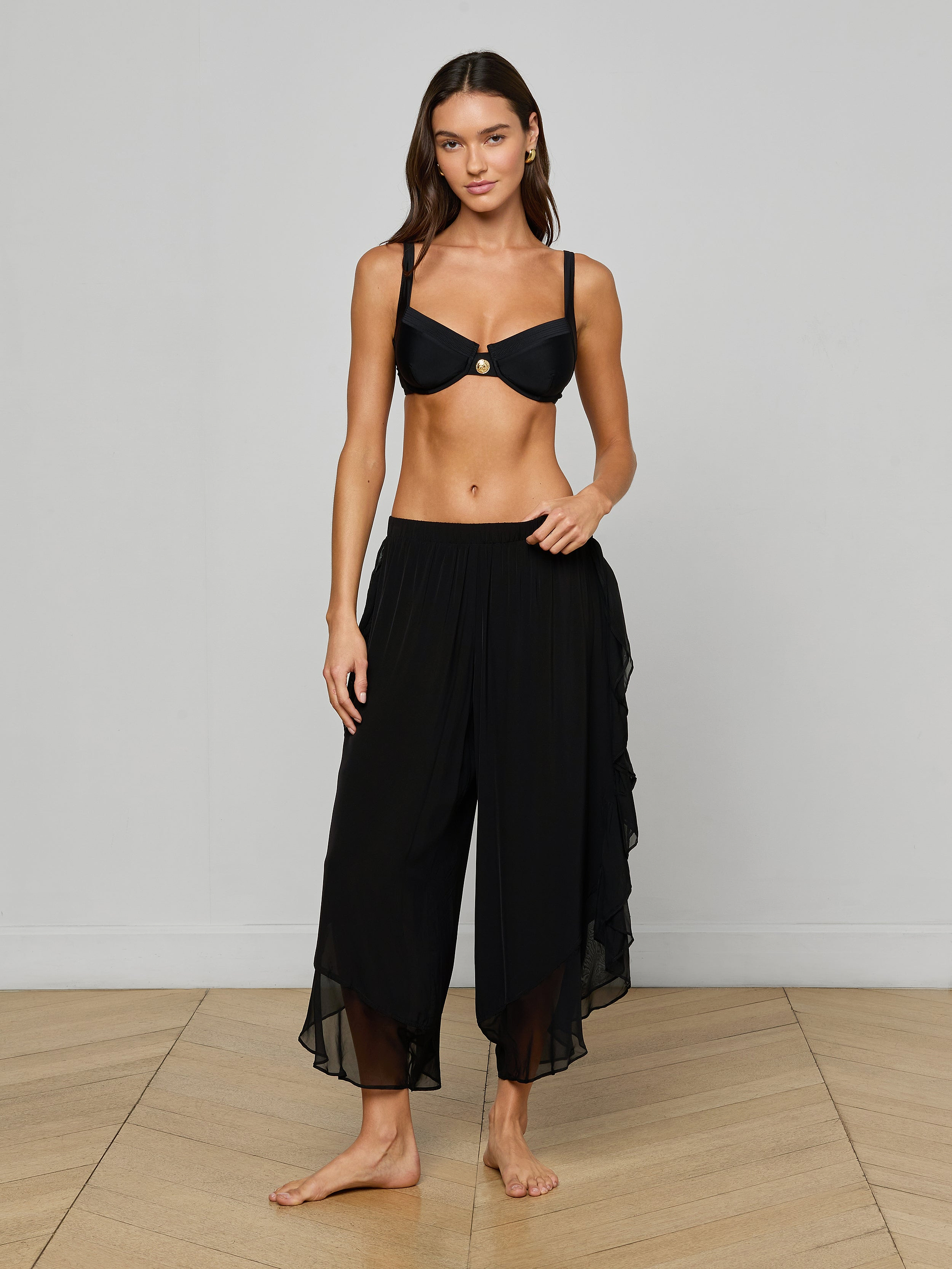 A woman stands barefoot against a plain light background, wearing a black bikini top and the Adonis Cover-Up Pant—loose, flowy pants made from sheer chiffon.