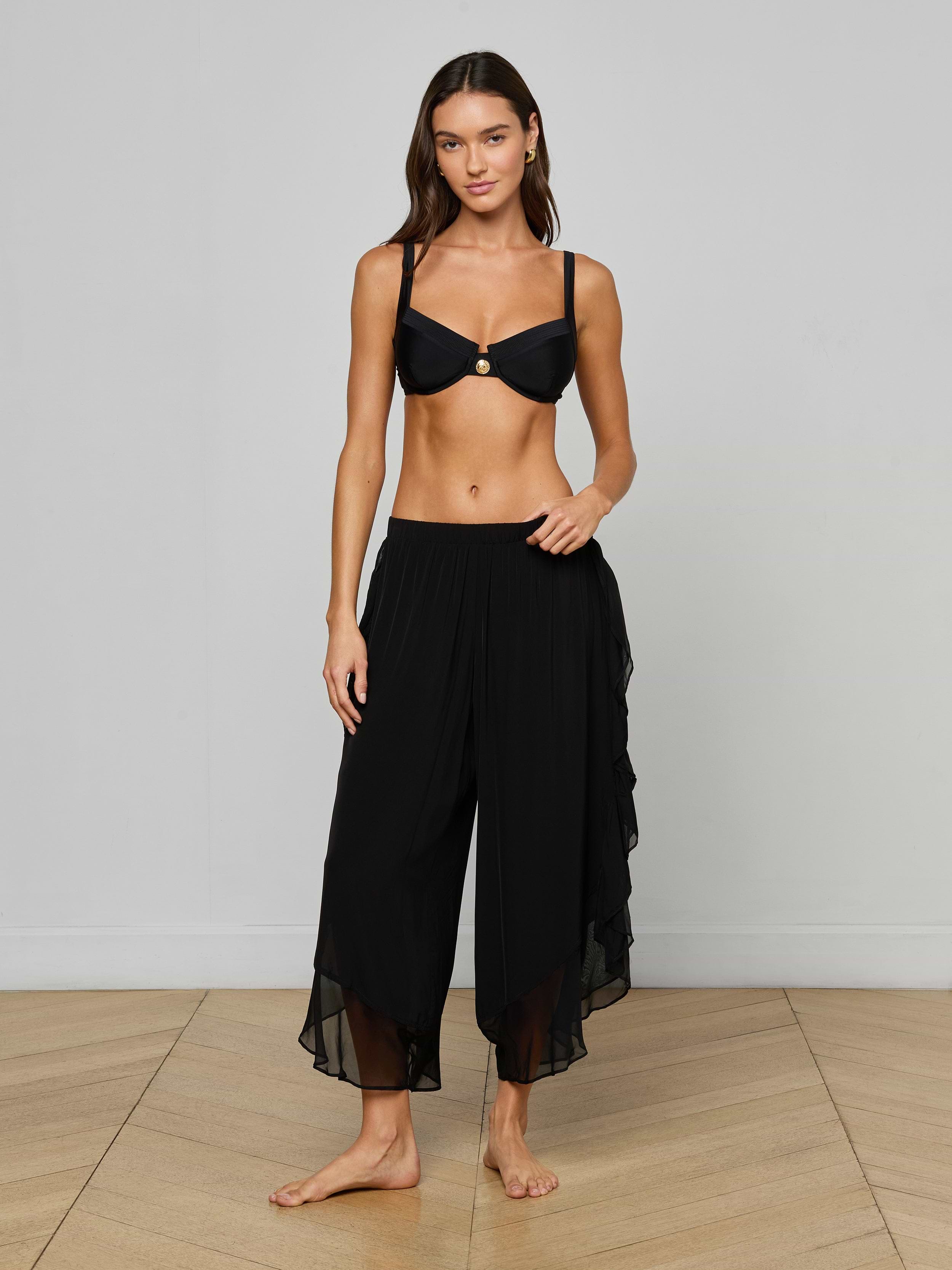 A woman stands barefoot against a plain light background, wearing a black bikini top and the Adonis Cover-Up Pant—loose, flowy pants made from sheer chiffon.