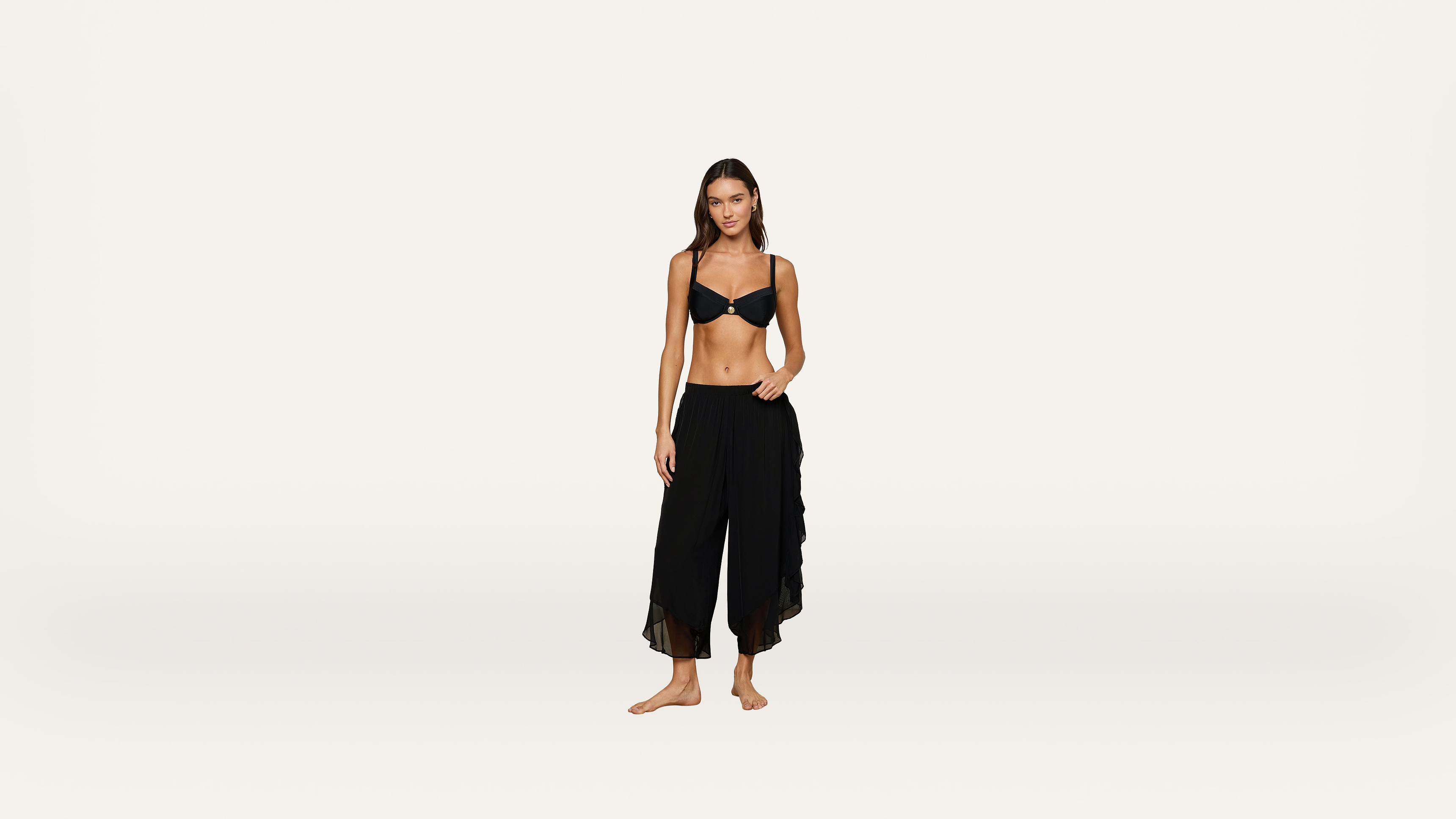 A woman stands barefoot against a plain white background, wearing a black bra and loose, sheer black pants.