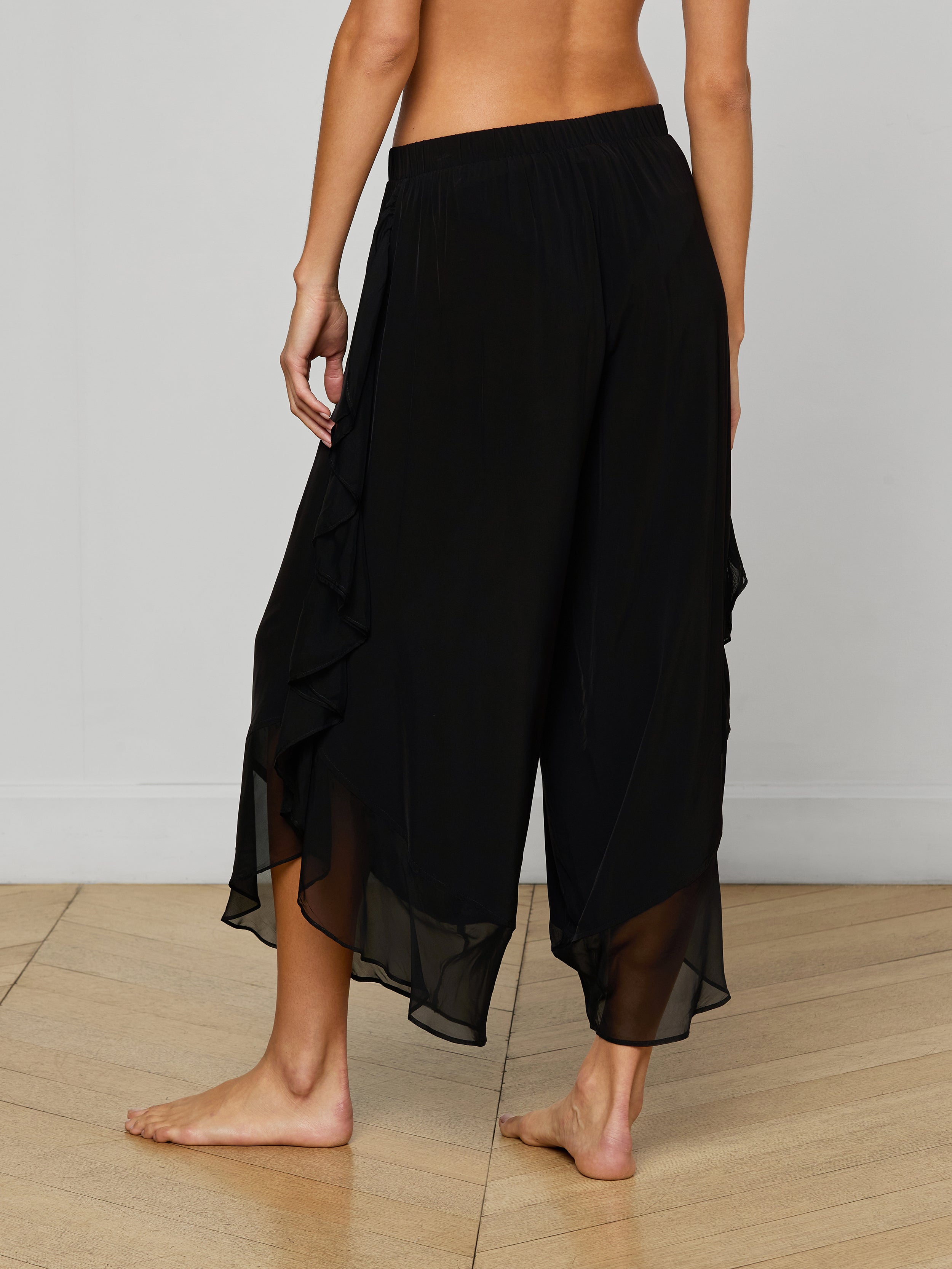 A person stands barefoot, showcasing the Adonis Cover-Up Pant—a pair of loose, black flowy pants with ruffled side details crafted from sheer chiffon. The image captures the wearer from the waist down against a plain light background, evoking an effortless poolside style.