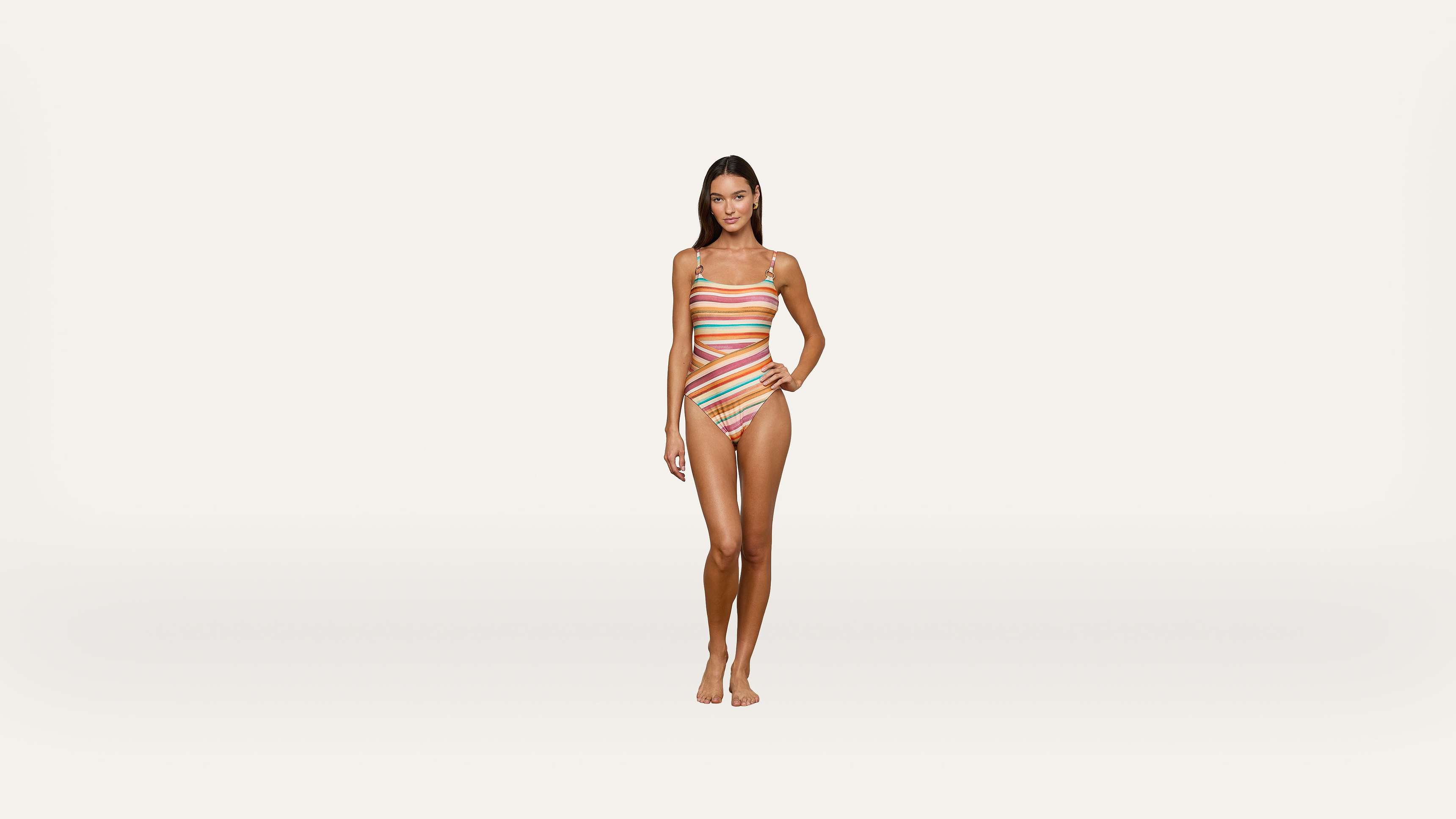 A woman stands facing forward, wearing a colorful, striped one-piece swimsuit with cutouts. She stands barefoot against a plain white background.