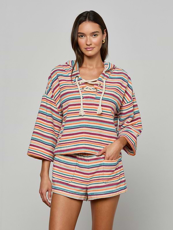 Model wears the Lee Cover-Up Poncho—a vibrant, striped, long-sleeve hooded top with a lace-up neckline and matching shorts—perfect for après-swim or poolside lounging. She stands facing forward with one hand in her shorts pocket against a plain background.