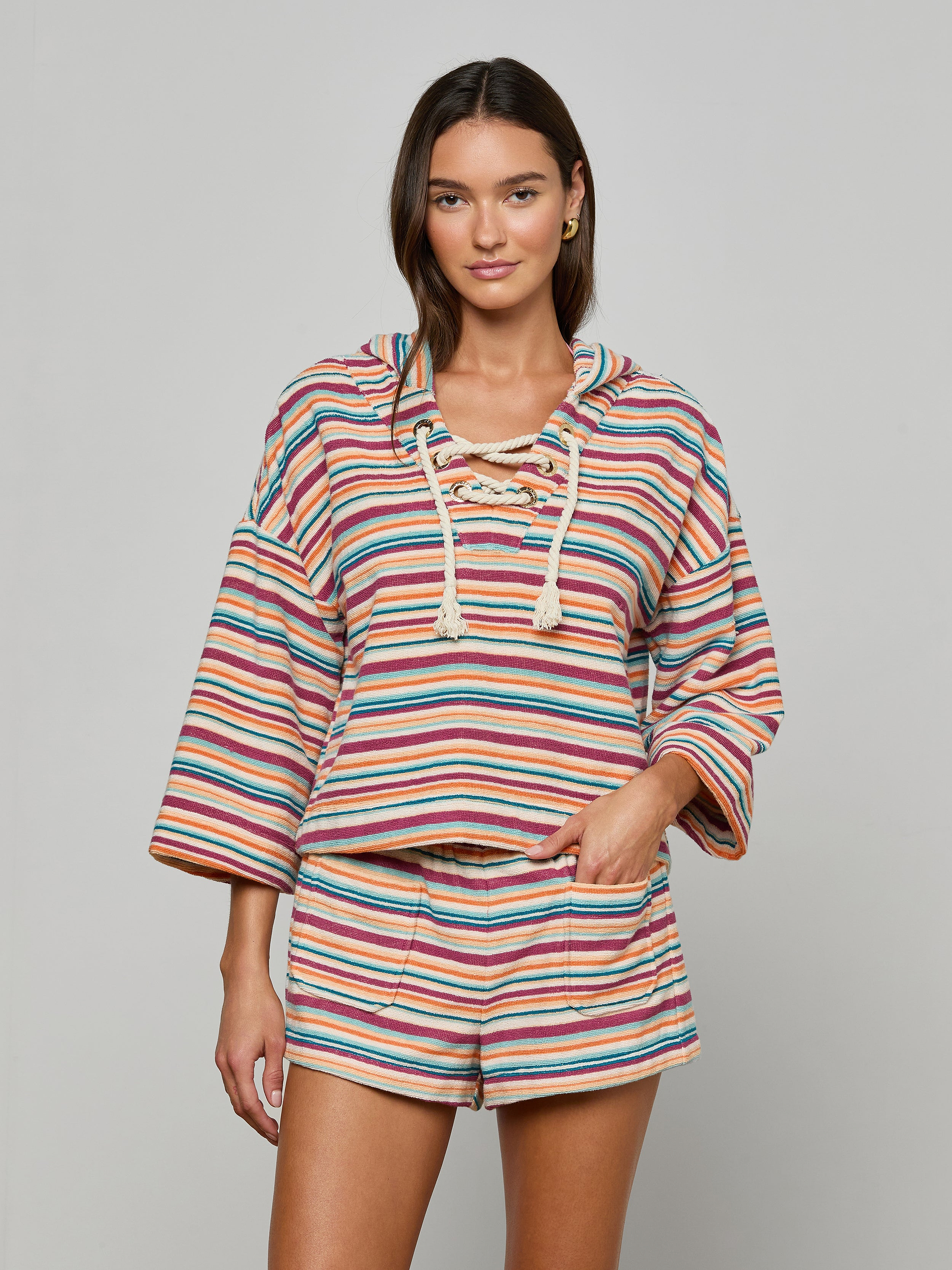 Model wears the Lee Cover-Up Poncho—a vibrant, striped, long-sleeve hooded top with a lace-up neckline and matching shorts—perfect for après-swim or poolside lounging. She stands facing forward with one hand in her shorts pocket against a plain background.