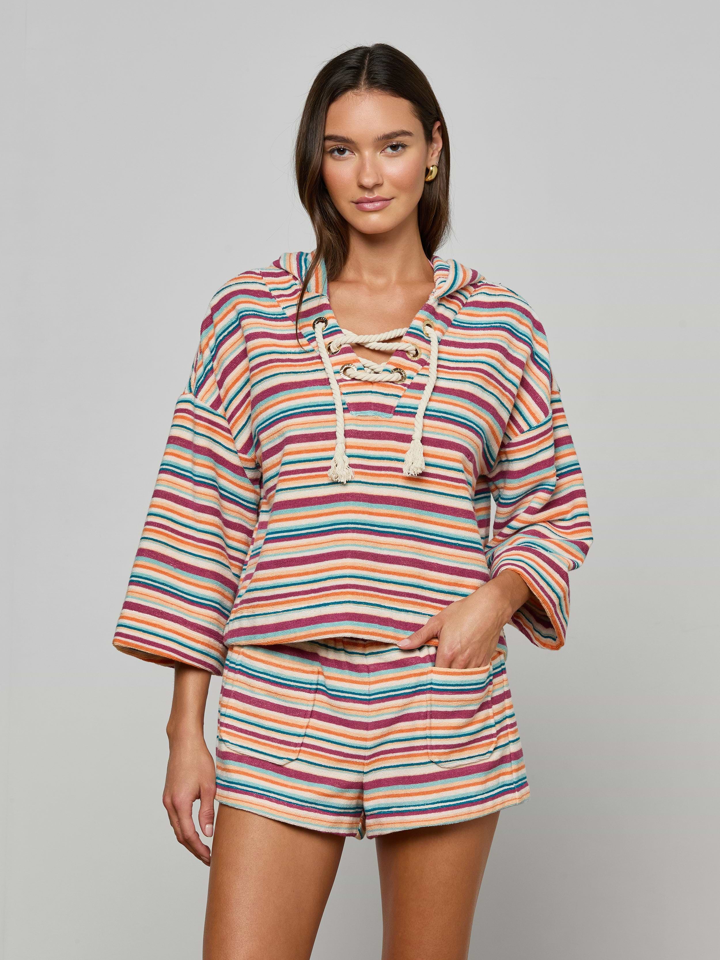 Model wears the Lee Cover-Up Poncho—a vibrant, striped, long-sleeve hooded top with a lace-up neckline and matching shorts—perfect for après-swim or poolside lounging. She stands facing forward with one hand in her shorts pocket against a plain background.
