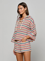 Wearing the Lee Cover-Up Poncho—a colorful, striped hoodie with a lace-up front and matching shorts—this woman stands with one hand in her pocket against a plain light background, ready for relaxed poolside lounging.