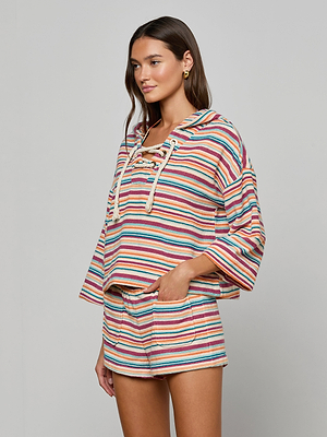 A woman stands against a plain light gray background wearing the Lee Cover-Up Poncho—a loose-fitting, long-sleeve, hooded top with matching multicolored striped shorts—ideal for poolside lounging or après-swim.
