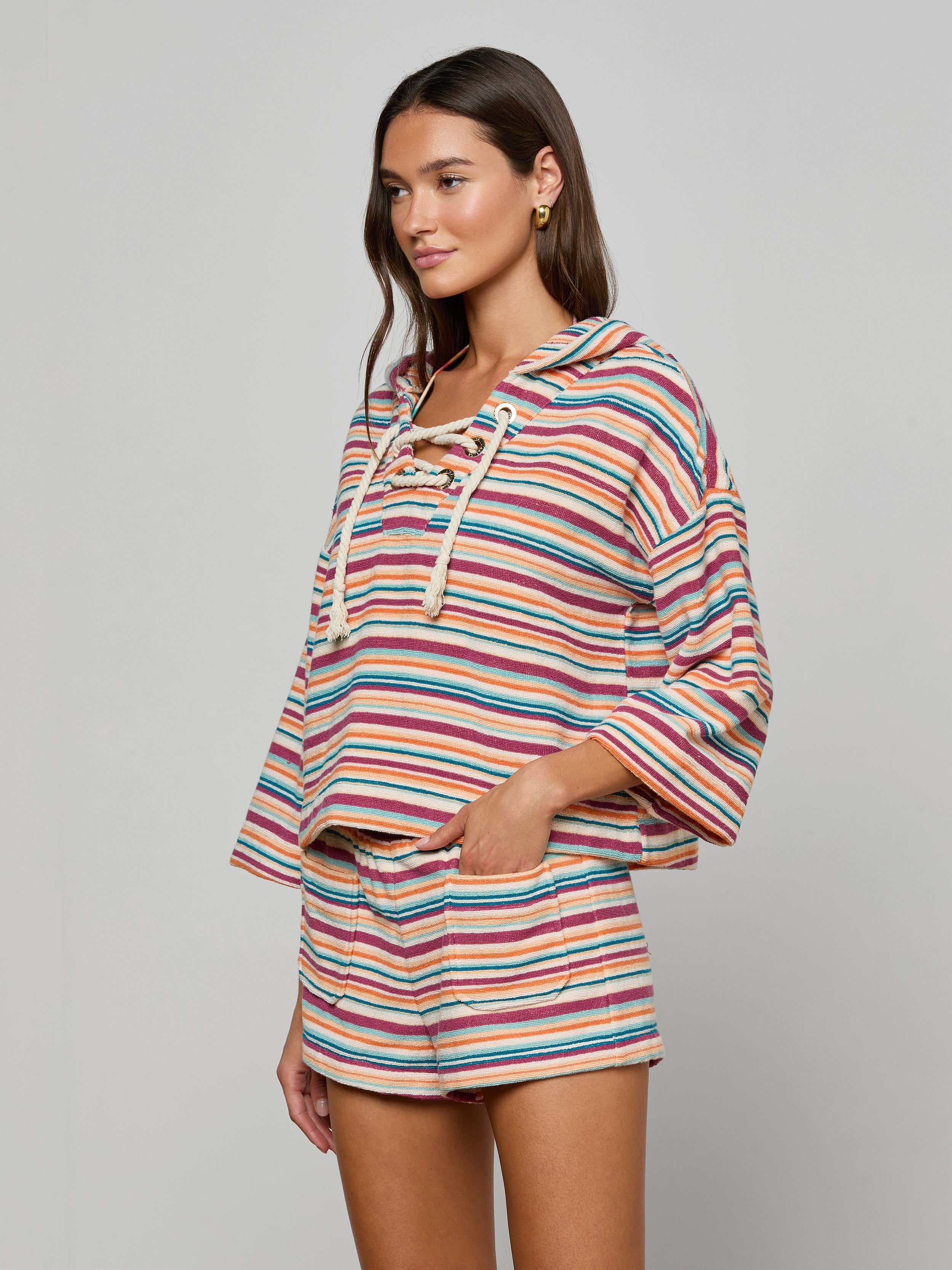 Wearing the Lee Cover-Up Poncho—a colorful, striped hoodie with a lace-up front and matching shorts—this woman stands with one hand in her pocket against a plain light background, ready for relaxed poolside lounging.