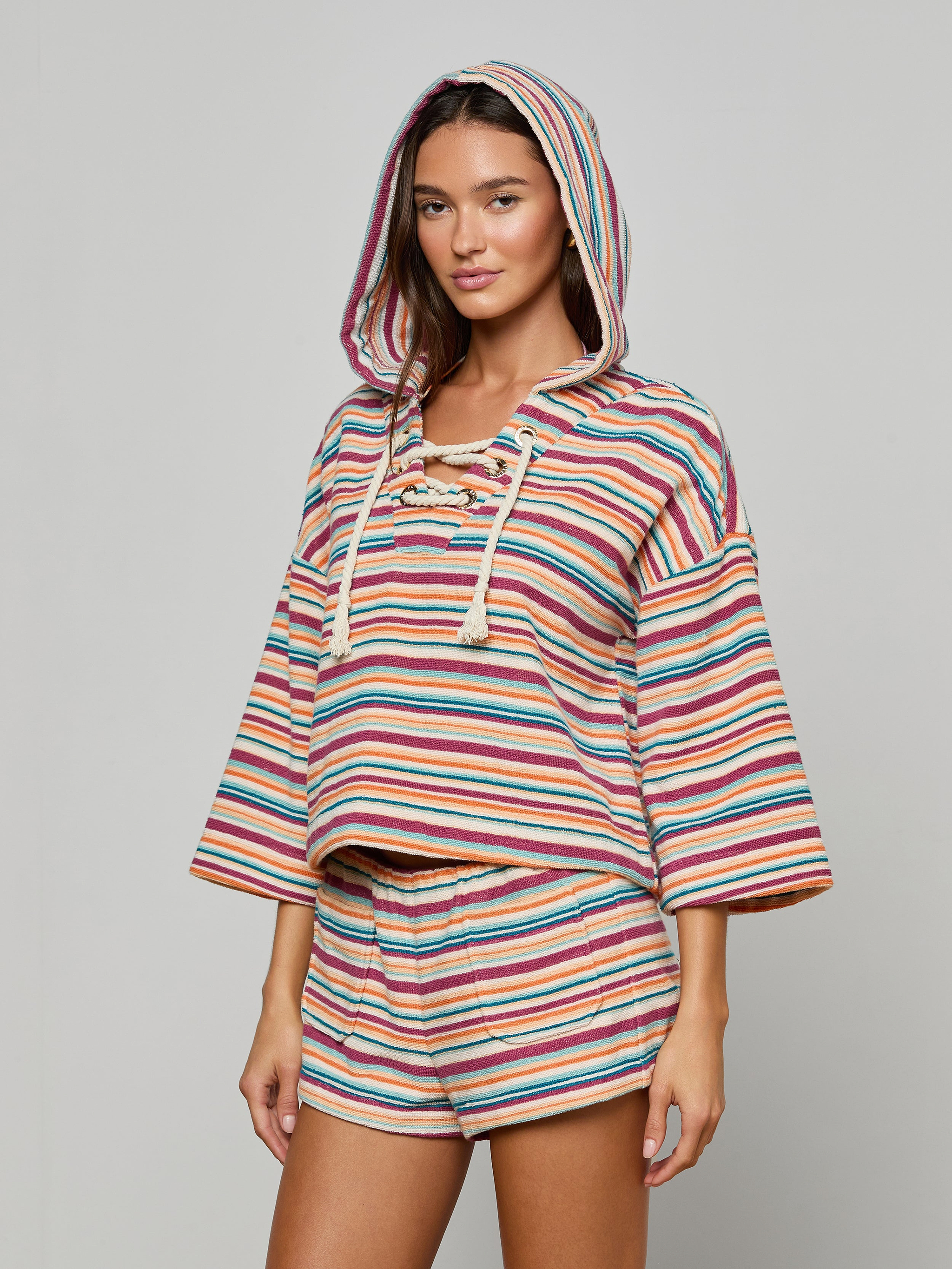 A woman wearing the Lee Cover-Up Poncho—a hooded, long-sleeve terrycloth poncho with matching shorts featuring colorful horizontal stripes—stands against a plain light background, making it an ideal choice for poolside lounging or après-swim relaxation.