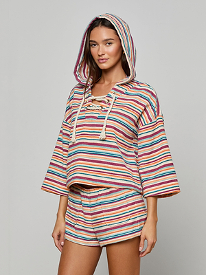 A woman stands against a plain light background wearing the Lee Cover-Up Poncho, a loose hooded terrycloth poncho with matching shorts featuring colorful horizontal stripes—perfect for poolside lounging.