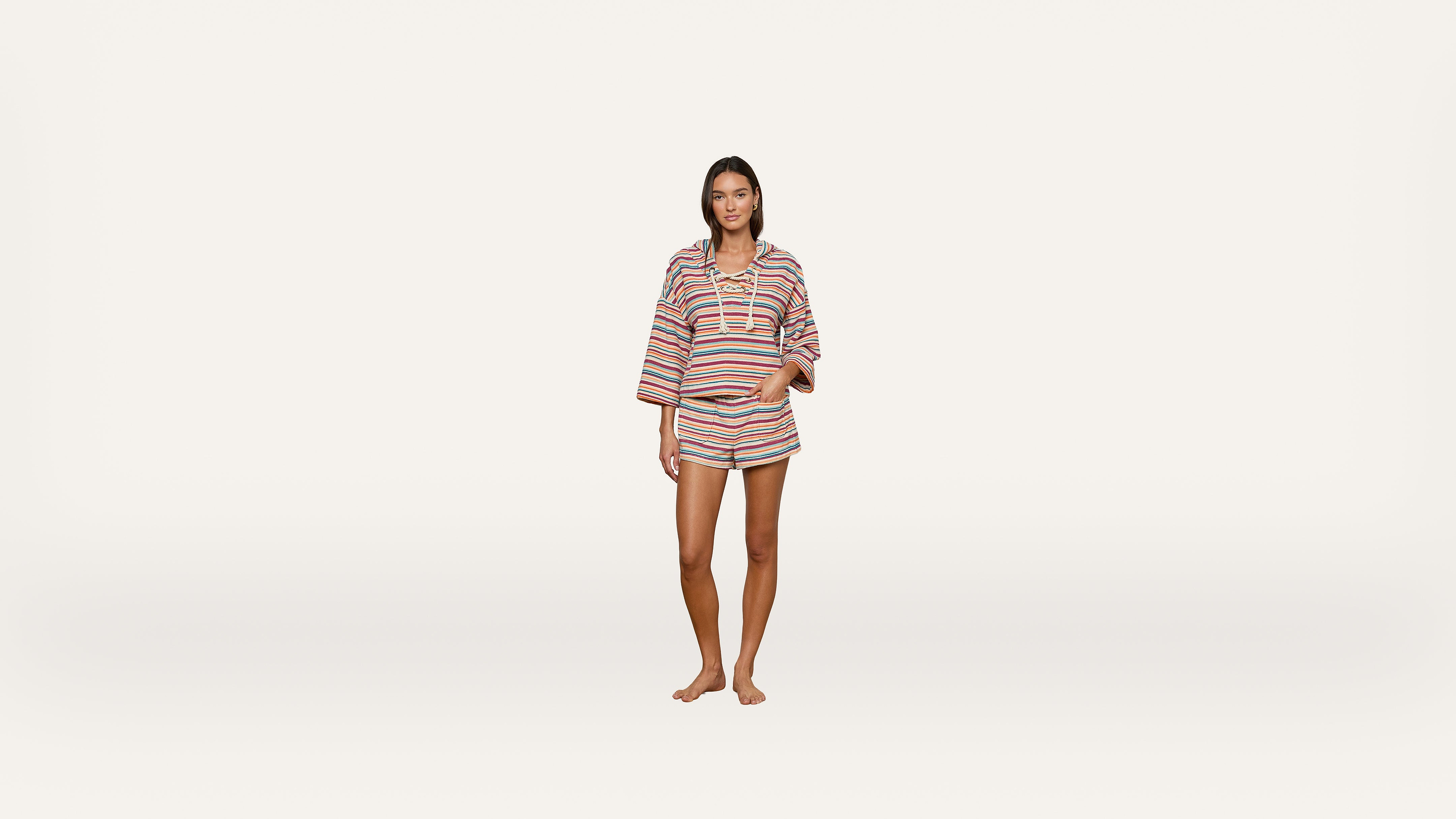 A woman stands barefoot against a white background, wearing a colorful, striped, long-sleeve top and matching shorts.