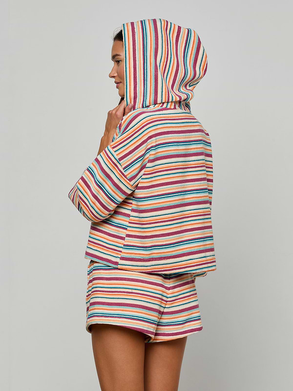 Woman in the Lee Cover-Up Poncho—a colorful, striped terrycloth hooded top with matching shorts—stands sideways against a plain background, her hood up and hand touching her chin. Perfect for après-swim or poolside lounging.