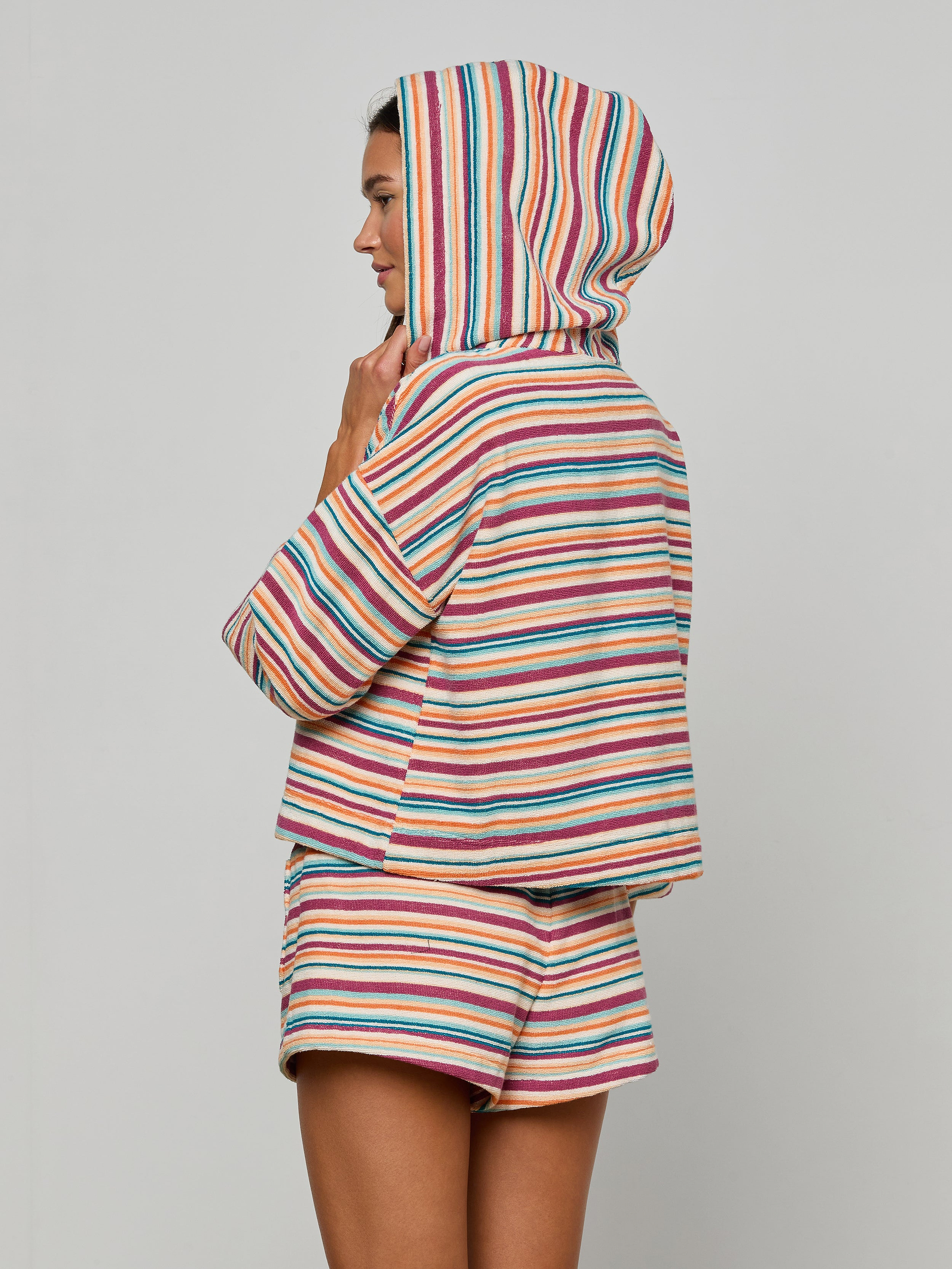 Woman in the Lee Cover-Up Poncho—a colorful, striped terrycloth hooded top with matching shorts—stands sideways against a plain background, her hood up and hand touching her chin. Perfect for après-swim or poolside lounging.