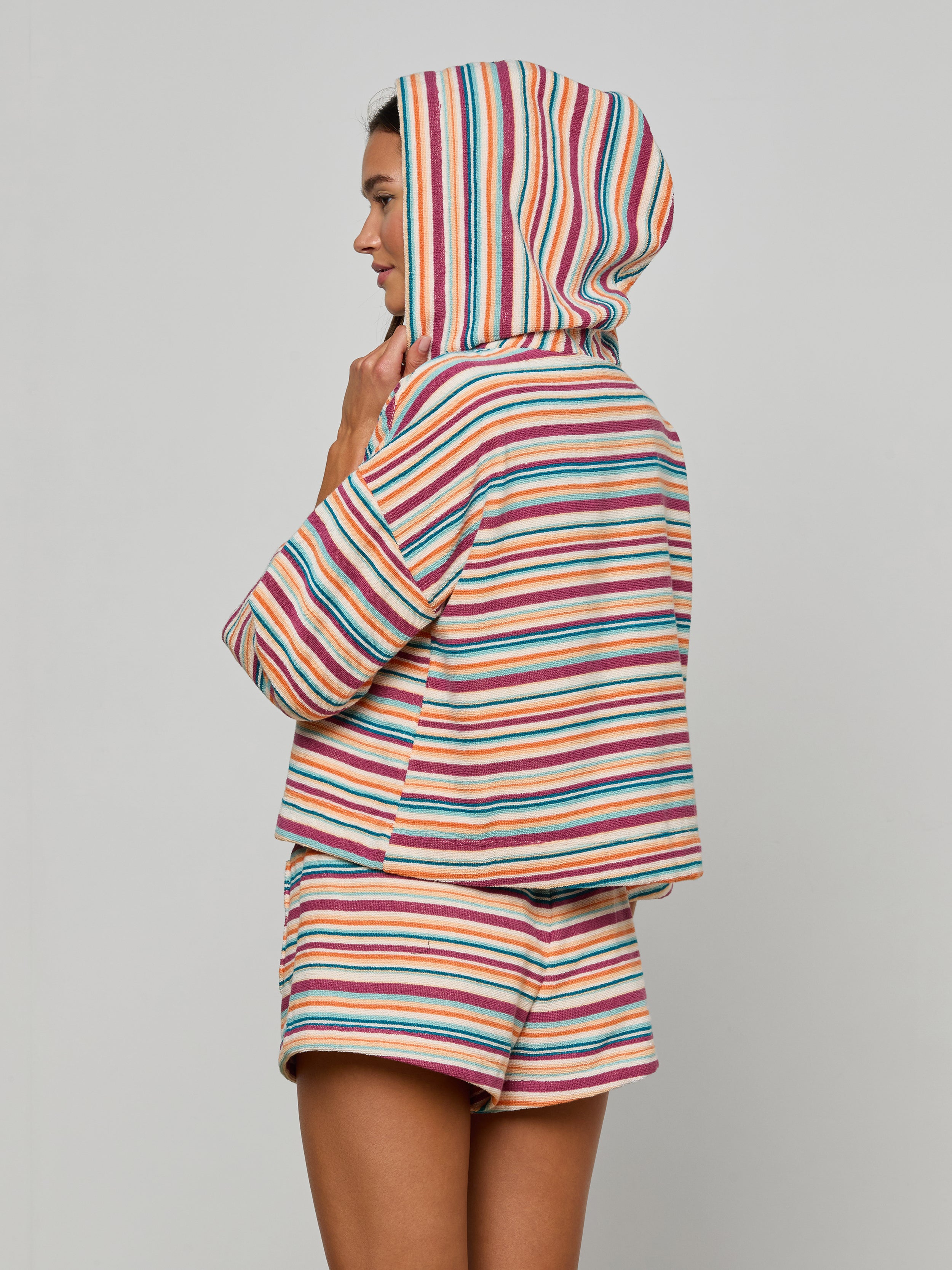 Woman in the Lee Cover-Up Poncho—a colorful, striped terrycloth hooded top with matching shorts—stands sideways against a plain background, her hood up and hand touching her chin. Perfect for après-swim or poolside lounging.