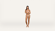 A woman stands barefoot against a plain white background, wearing a striped bikini with her arms folded.