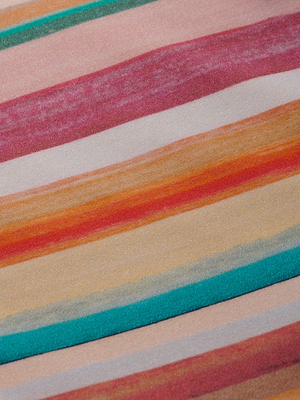 A close-up of the Nicole Bikini Bottom's four-way stretch nylon shows visible texture and vibrant diagonal watercolor stripes in pink, red, orange, yellow, teal, and beige.