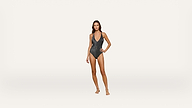 Woman standing barefoot against a plain white background, wearing a dark, textured one-piece swimsuit with a deep V-neck and thin shoulder straps. Her right hand rests on her hip and she looks straight ahead.
