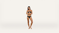 A woman stands barefoot against a plain white background, wearing a black bikini with a decorative strap detail, her arms crossed over her midsection.
