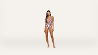 A woman stands barefoot against a plain white background, wearing a floral one-piece swimsuit with a plunging neckline and halter straps.