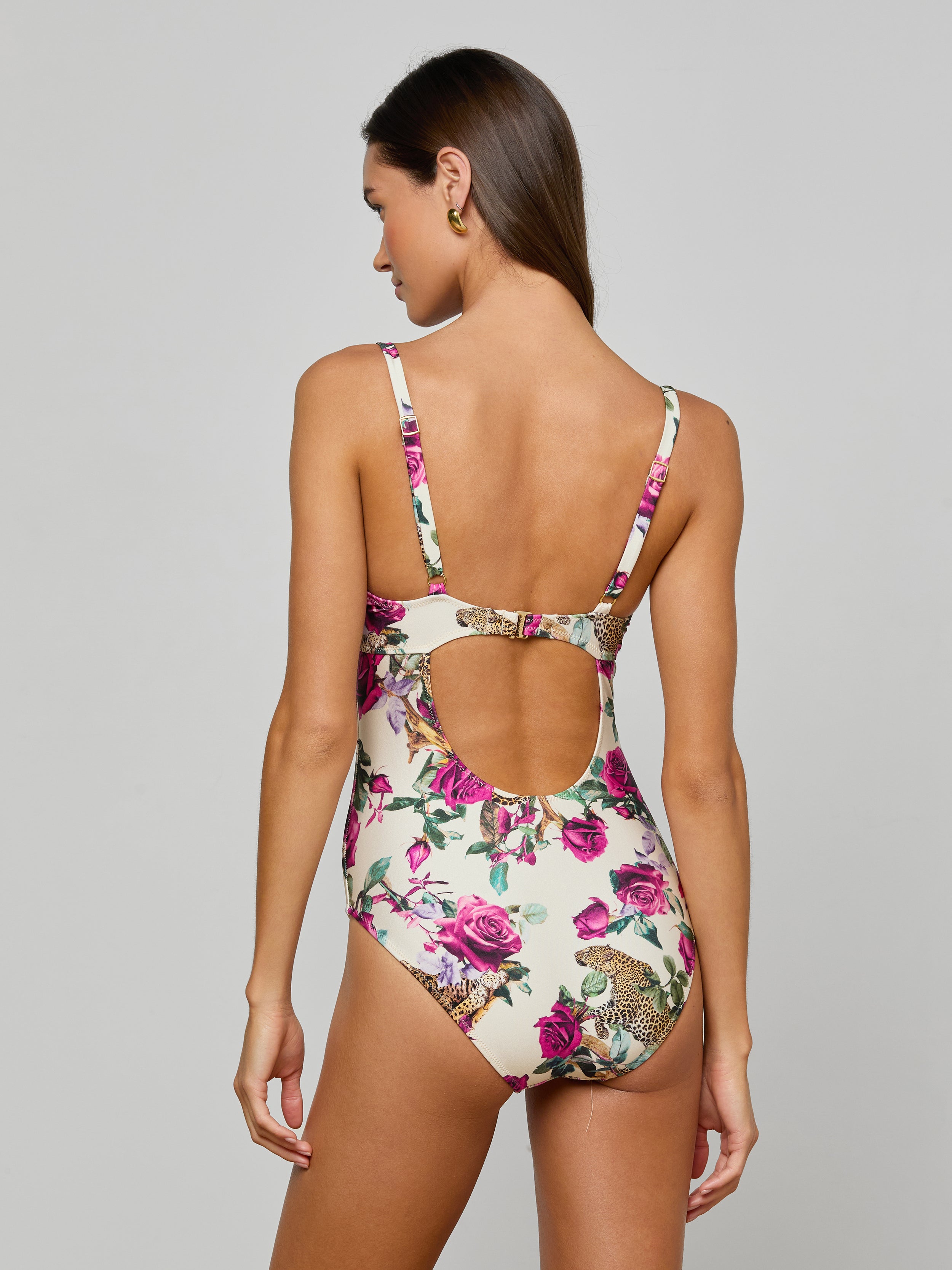 A woman faces away, wearing the Amie Bandeau One-Piece Swimsuit with a leopard print and pink floral accents. This swimsuit features a low scoop back with a circular cutout and provides UPF 50 sun protection. The background is plain and light-colored.