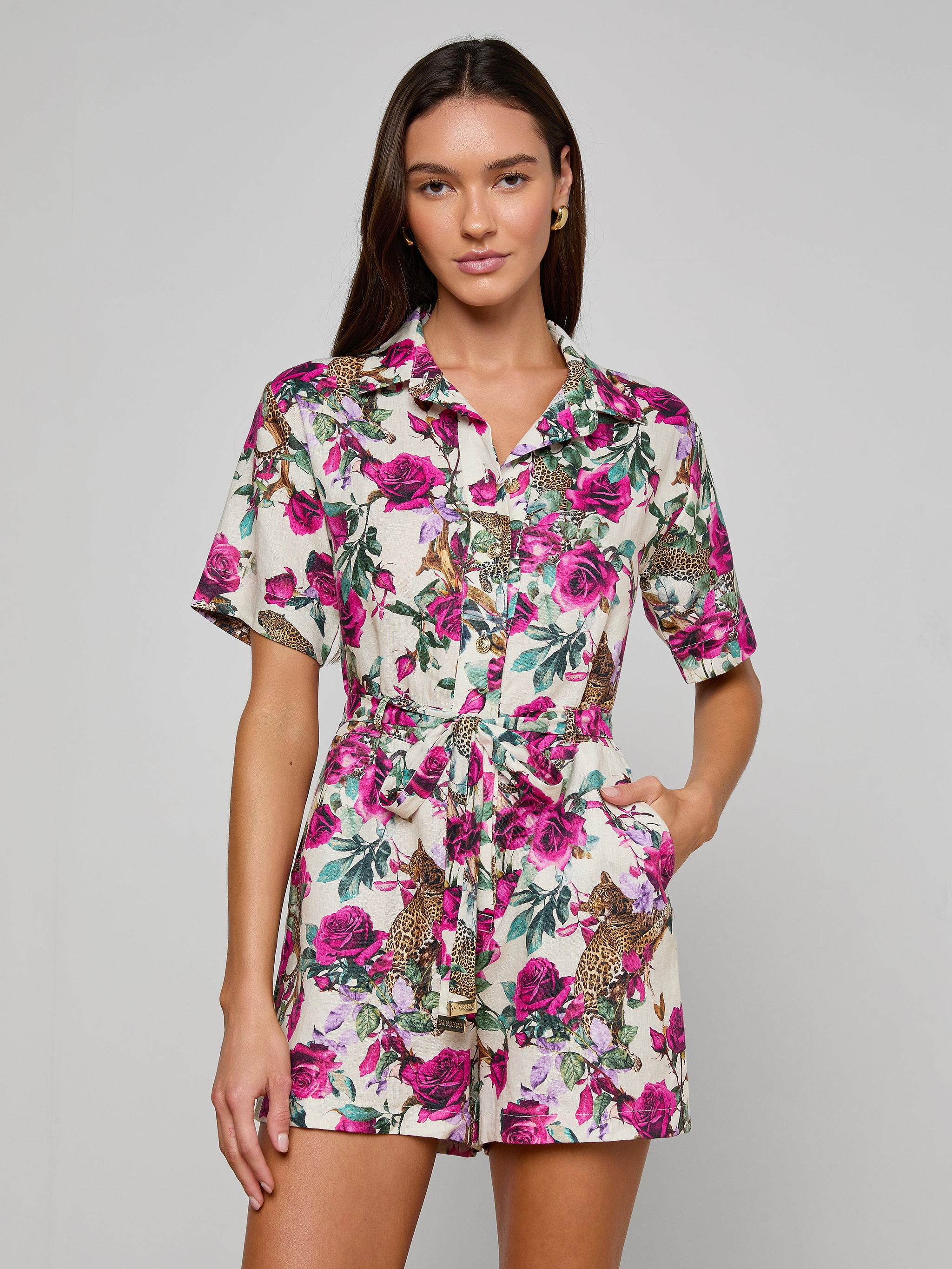 A woman stands with her hand in her pocket, facing forward against a plain light background, wearing the Hestia Cover-Up Romper—a short-sleeved, button-up linen romper featuring a pink floral and leopard print pattern.
