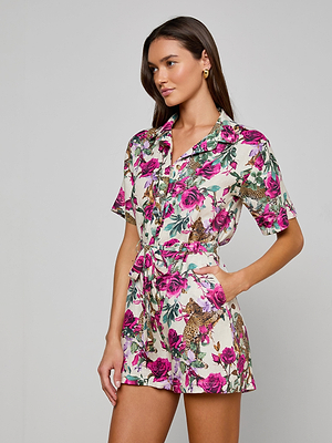 A woman poses in the Hestia Cover-Up Romper, a short-sleeve, button-up linen piece with pink, green, and beige floral and animal prints. Features include waist tie and pockets, inspired by rose leopard print swimwear.