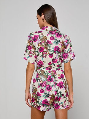 A woman faces away, wearing the Hestia Cover-Up Romper—a short-sleeved linen piece with vibrant pink roses and leopards on white, a collar, and gold hoop earrings—standing out against a plain gray background.