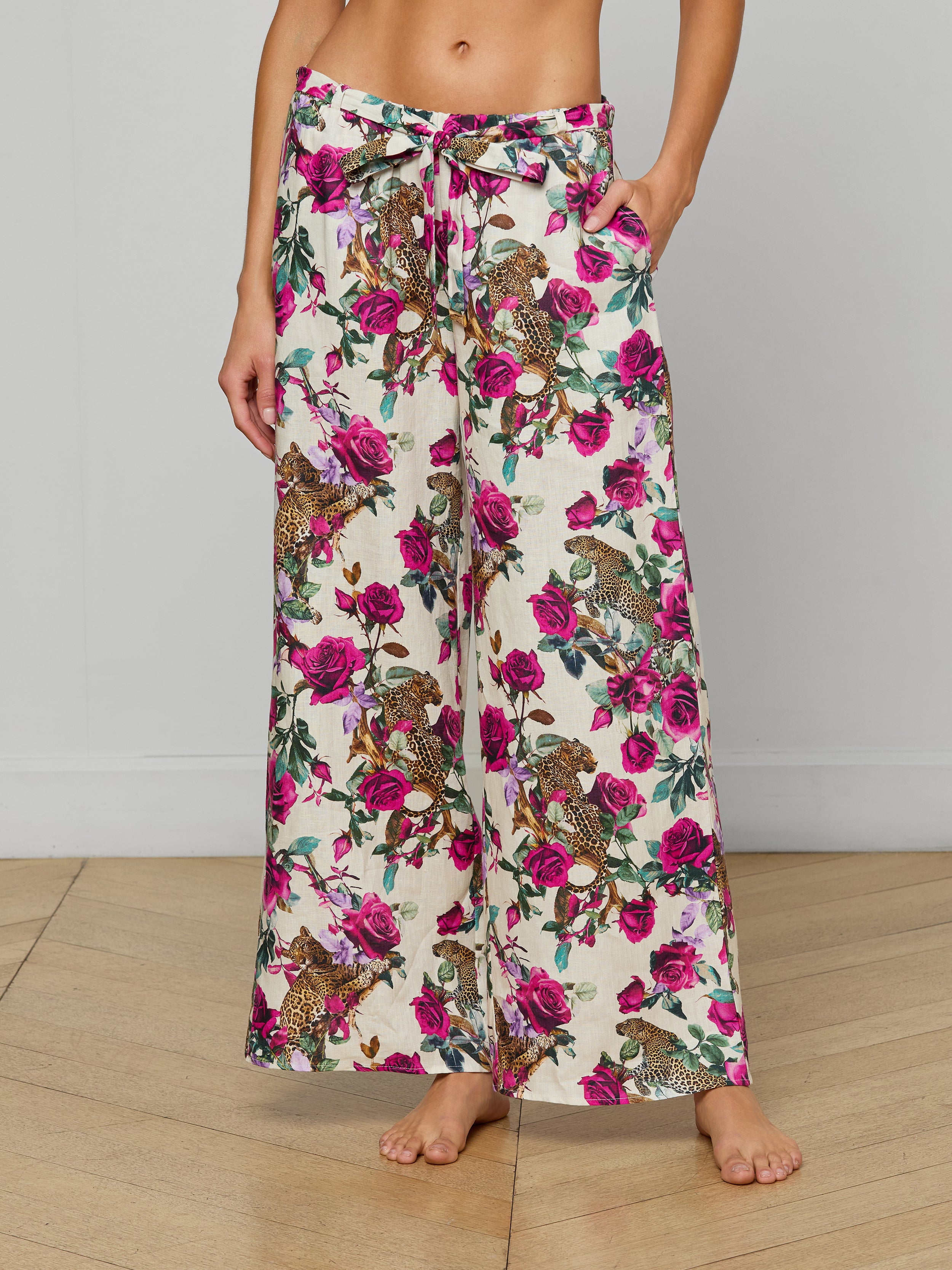 A woman models the Geraldine Cover-Up Pant, a wide-leg, high-waisted linen resort style featuring a pink and green rose leopard print. The pants include a tie waist and side pockets. She stands barefoot against a plain background.